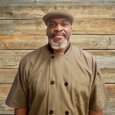 A man with a gray beard wearing a green chef coat and cap stands in front of a wooden plank wall, proudly representing Thrive Senior Living.