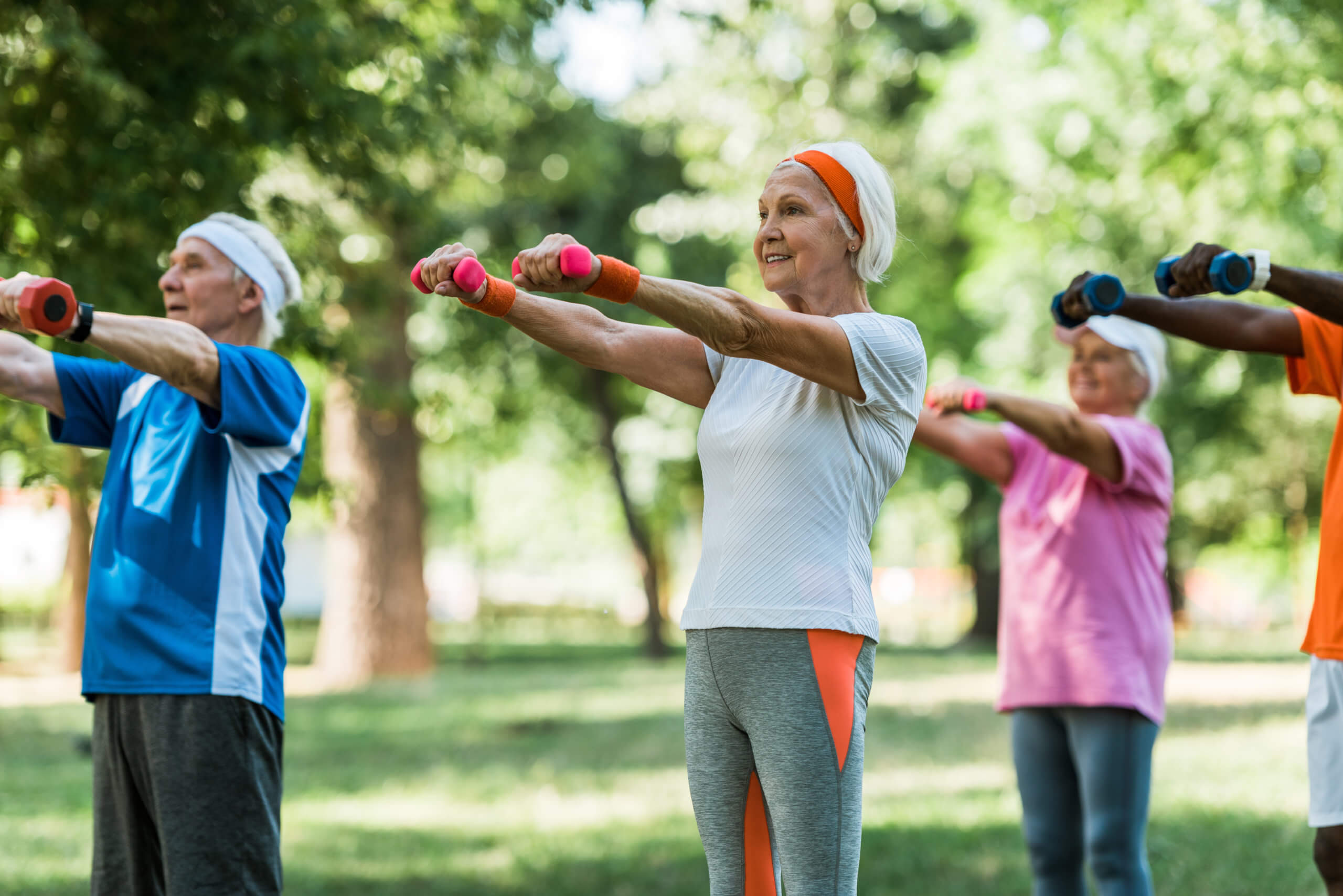 A group of older adults at Thrive Senior Living exercises outdoors, standing in a row holding small dumbbells with arms extended, surrounded by trees and greenery.