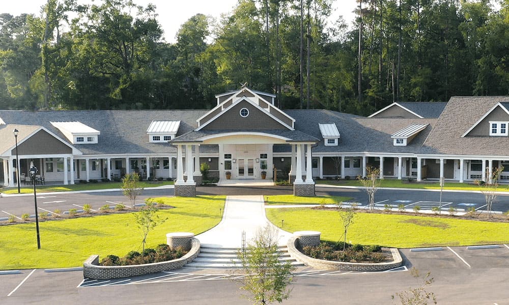 A single-story Thrive Senior Living building with a covered entrance, white pillars, and a landscaped front lawn, surrounded by trees and an empty parking lot.
