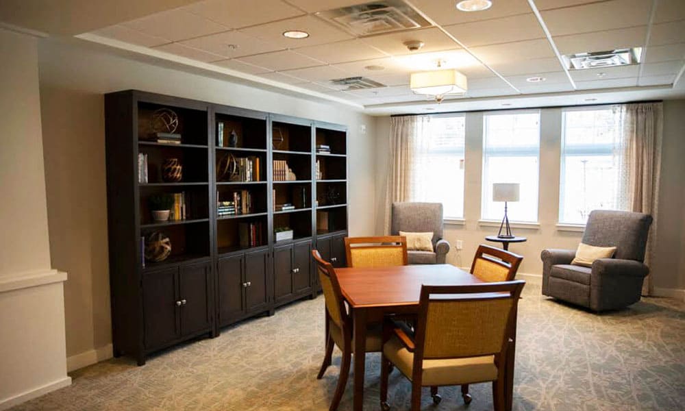 A cozy room at Thrive Senior Living with a wooden table and four chairs, two armchairs by large windows, a floor lamp, and a tall bookshelf filled with books and decor.