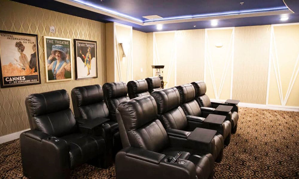 Home theater room at Thrive Senior Living with rows of black leather recliners, patterned carpet, vintage movie posters on the wall, and soft yellow lighting.