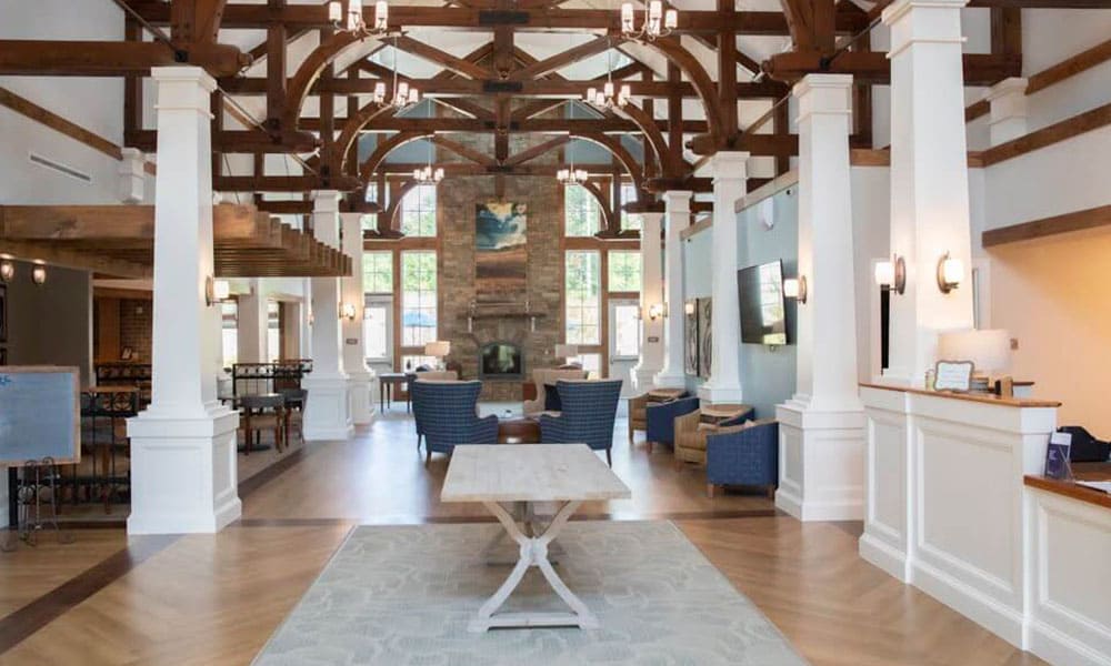 Spacious lobby at Thrive Senior Living with exposed wooden beams, white columns, a stone fireplace, blue armchairs, and large windows allowing natural light.