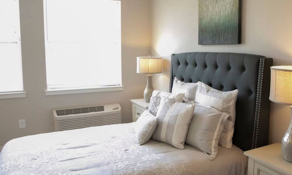 A neatly made bed with multiple pillows sits between two nightstands with lamps in a bright Thrive Senior Living bedroom featuring two windows and a wall-mounted air conditioner.