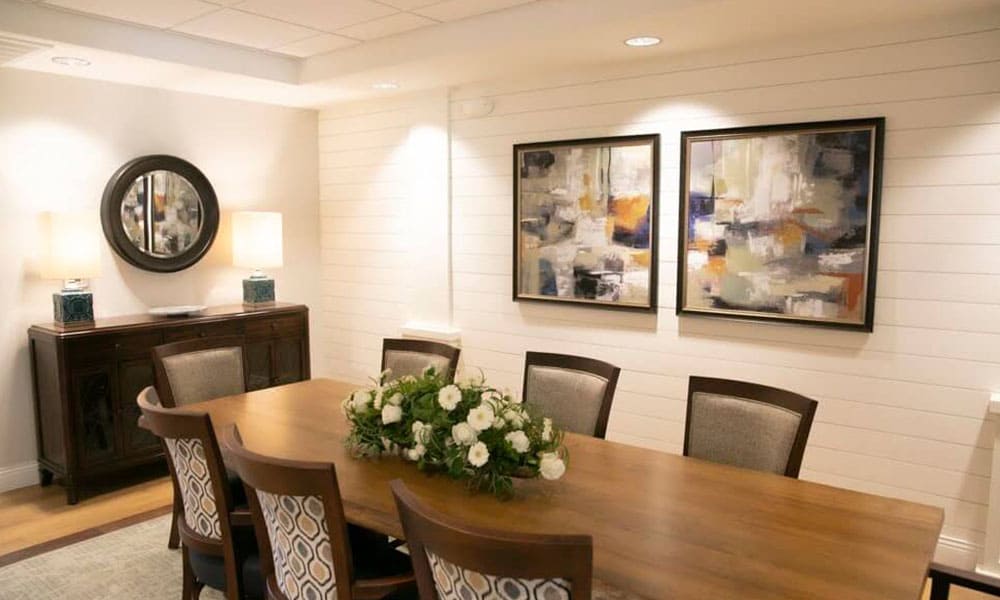 A dining room at Thrive Senior Living features a wooden table, eight chairs, a floral centerpiece, two abstract paintings on the wall, and a sideboard with a round mirror and two lamps.