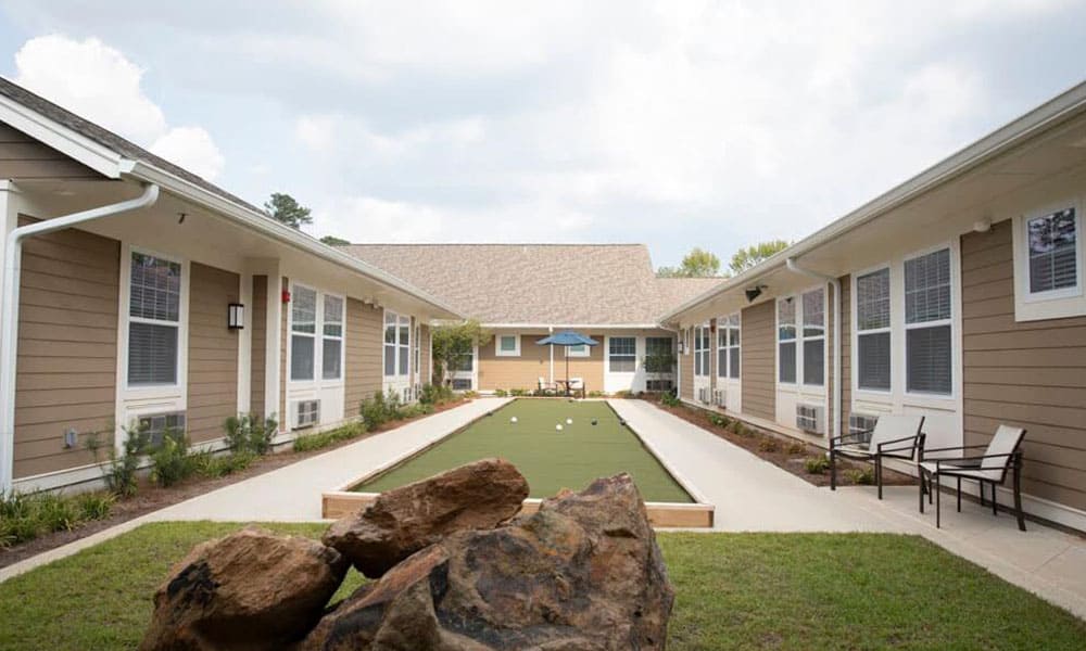 A rectangular bocce ball court is situated between two single-story tan buildings with white trim at Thrive Senior Living, surrounded by landscaped paths and outdoor seating.