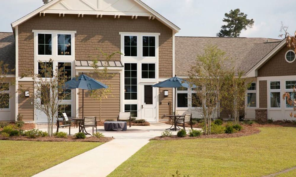 A modern two-story Thrive Senior Living building with large windows, outdoor seating areas with tables and blue umbrellas, and a landscaped lawn.