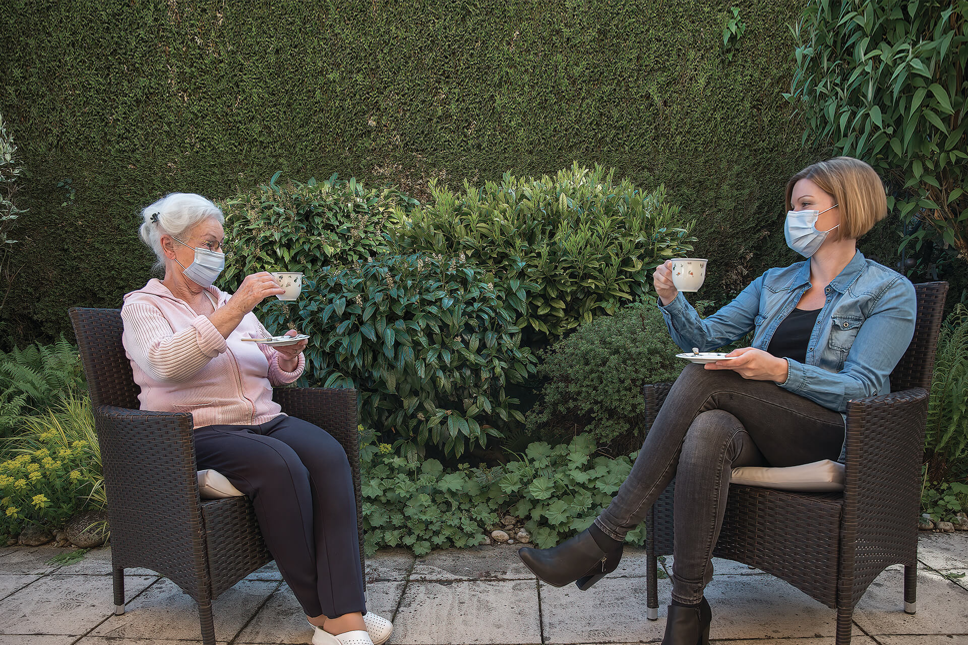Two women at Thrive Senior Living sit in wicker chairs outdoors, wearing face masks and holding teacups, with lush green foliage in the background.