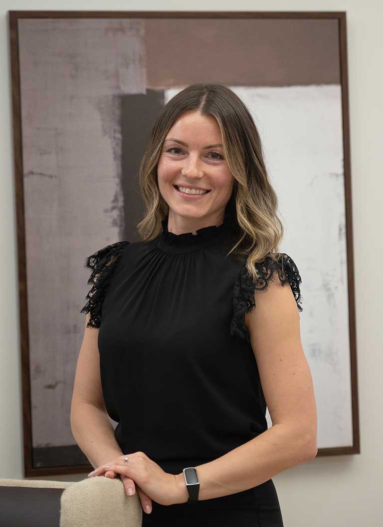 A woman with shoulder-length brown hair, wearing a black blouse and a smartwatch, stands indoors at Thrive Senior Living in front of a modern, abstract painting, smiling at the camera.