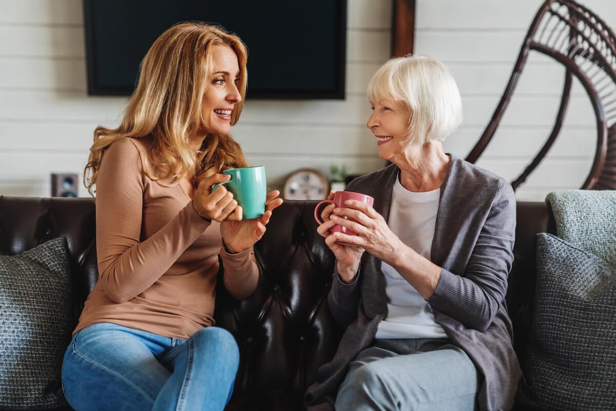 Two women sit on a couch at Thrive Senior Living, holding mugs and smiling at each other in a cozy living room setting.