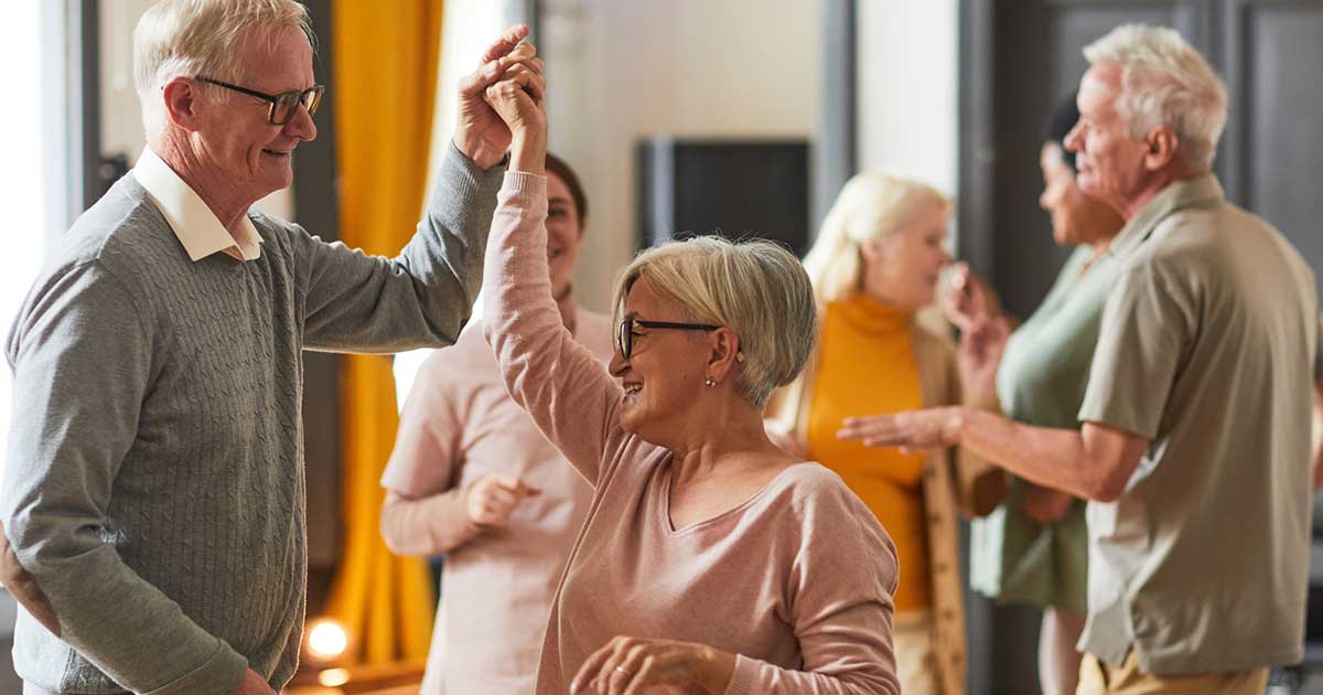 A group of older adults at Thrive Senior Living socialize, with two pairs dancing and others talking and smiling in a well-lit indoor setting.