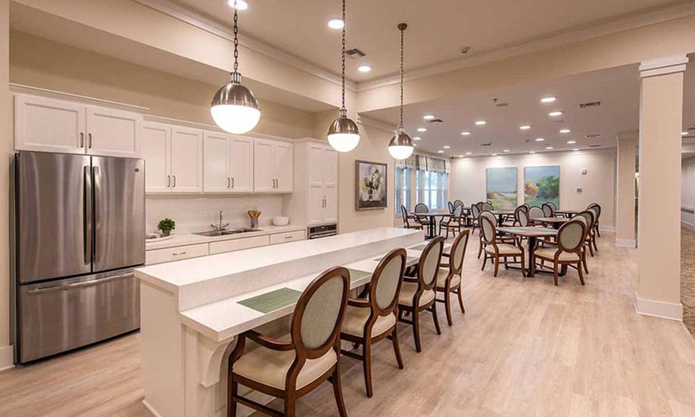 Spacious communal kitchen and dining area at Thrive Senior Living, featuring white cabinetry, stainless steel appliances, round dining tables, and upholstered chairs under elegant pendant lights.