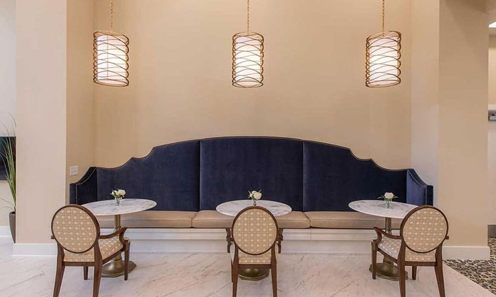 Three round tables with beige chairs and a long blue upholstered bench are set beneath three pendant lights in a modern, neutral-toned dining area at Thrive Senior Living.