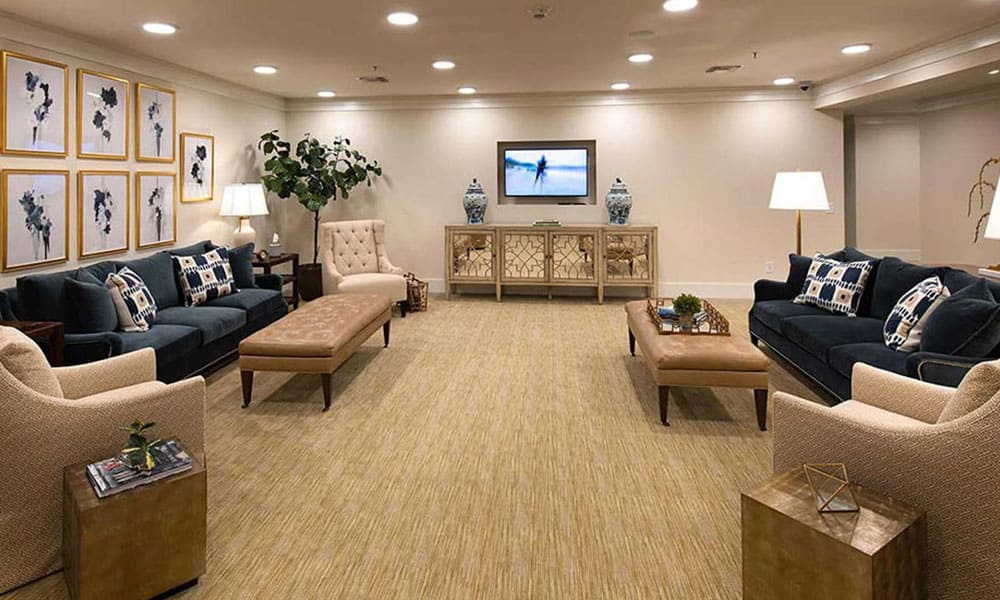 A spacious, well-lit living room at Thrive Senior Living with two dark sofas, beige armchairs, a bench, side tables, and a TV mounted on the wall above a console. Decorative art and lush plants create an inviting atmosphere.