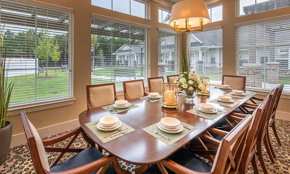 A dining room at Thrive Senior Living features a large wooden table set for eight, surrounded by chairs, with expansive windows offering lovely views of houses and greenery outside.