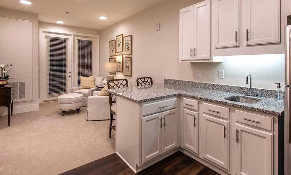 A modern open-plan kitchen with white cabinets, granite countertops, and a small sink opens to a cozy living area with beige furniture and framed art—the perfect space for Thrive Senior Living comfort and community.