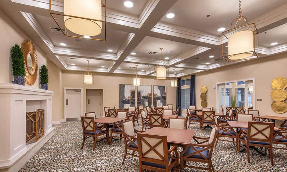Spacious dining room at Thrive Senior Living features square wooden tables, cushioned chairs, patterned carpet, pendant lights, and decorative wall art.