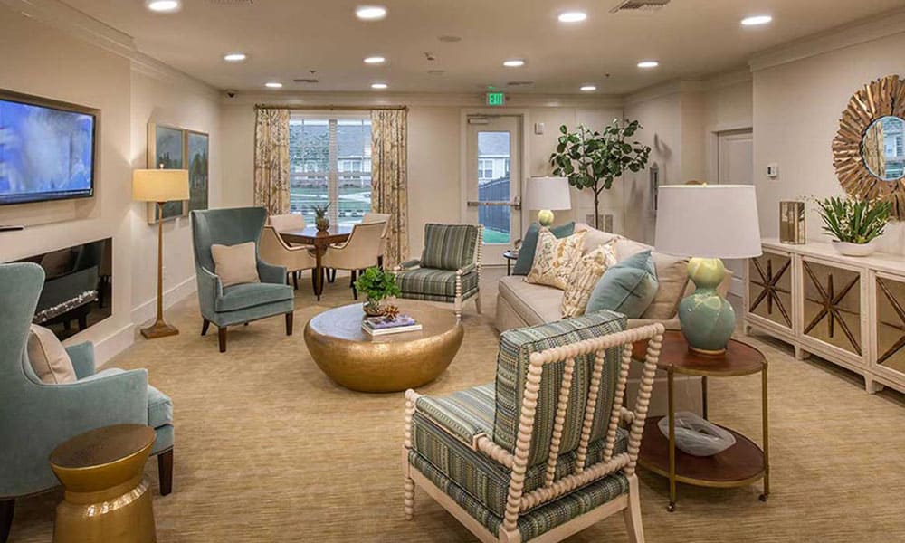 A spacious, well-lit living room at Thrive Senior Living with modern furniture, including armchairs, a round coffee table, lamps, decorative plants, and abstract wall art.