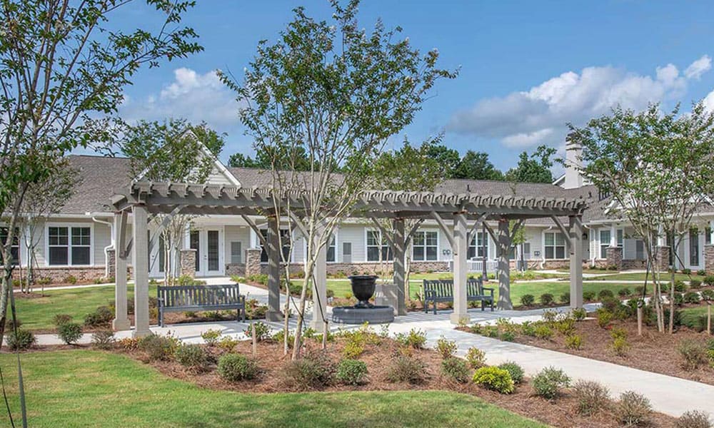 A landscaped courtyard at Thrive Senior Living features a pergola, benches, a central fountain, and a pathway leading to inviting single-story residential buildings.