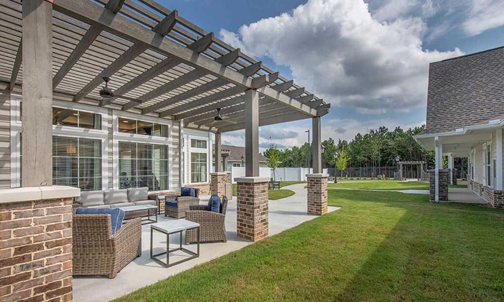 Outdoor patio with wicker furniture and tables under a pergola, adjacent to a brick building at Thrive Senior Living, overlooking a lawn and trees beneath a partly cloudy sky.
