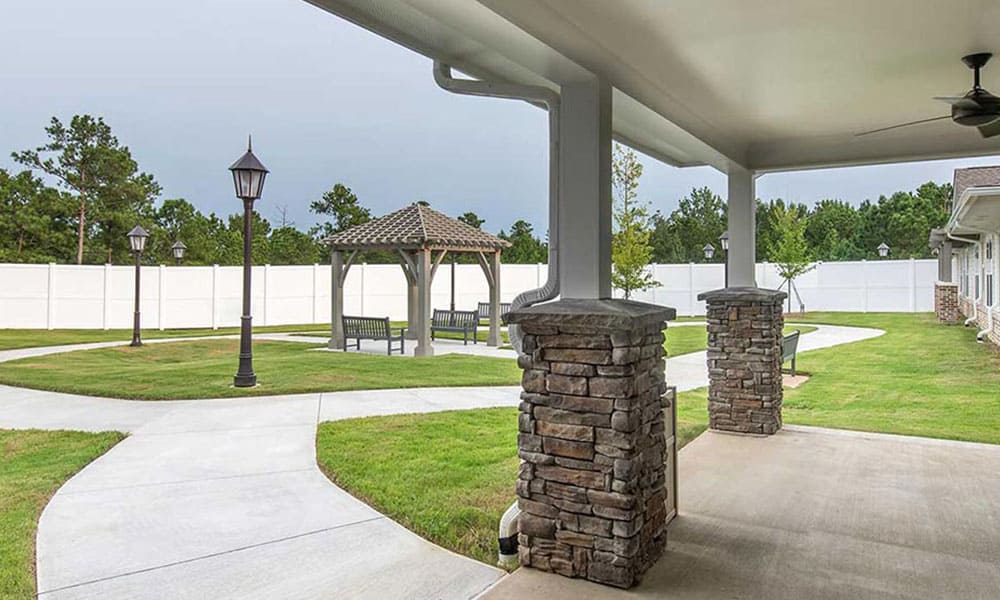 Covered patio with stone pillars overlooks a landscaped yard with winding walkways, lamp posts, benches, gazebo, and white privacy fence—an inviting outdoor retreat at Thrive Senior Living.