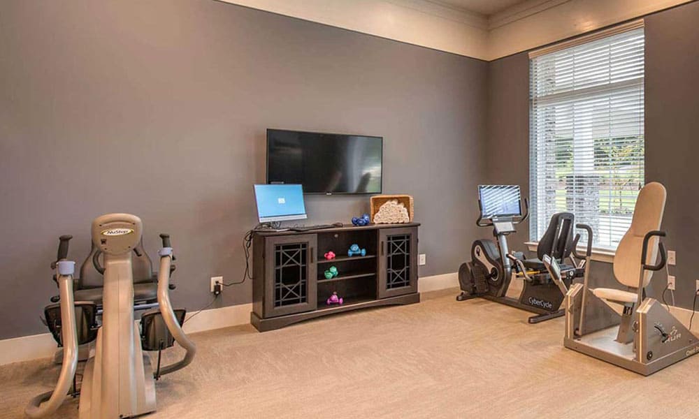 A small gym room at Thrive Senior Living features two exercise bikes, a flatscreen TV, a laptop, and a black console holding colorful dumbbells, all situated next to a large window with blinds.