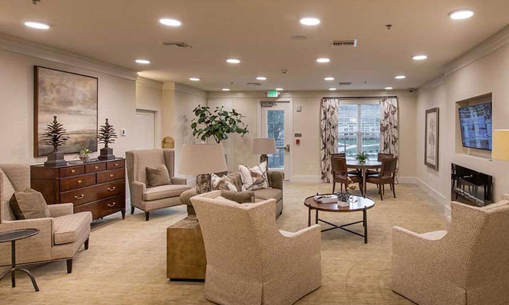 A well-lit lounge at Thrive Senior Living features beige armchairs, a sofa, a dresser, coffee tables, lush plants, wall art, and large windows with elegant drapes.