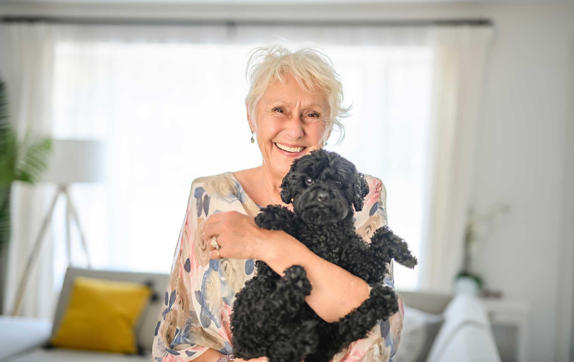 An older woman with short white hair smiles while holding a small black curly-haired dog in a bright living room at Thrive Senior Living.