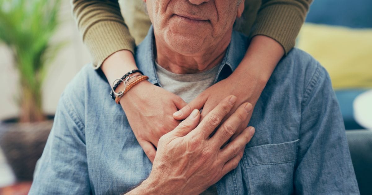 An older man sits while someone stands behind him, resting their arms gently around his shoulders and chest—a touching moment that reflects the caring atmosphere at Thrive Senior Living.