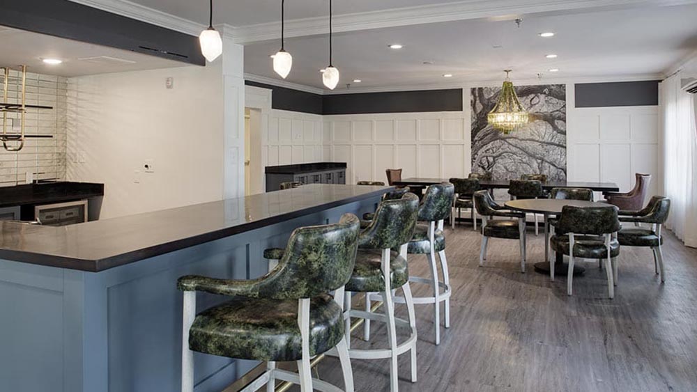 A modern lounge area at Thrive Senior Living features a bar counter, green upholstered chairs, round tables, and a chandelier, all set against wood flooring and panelled walls.