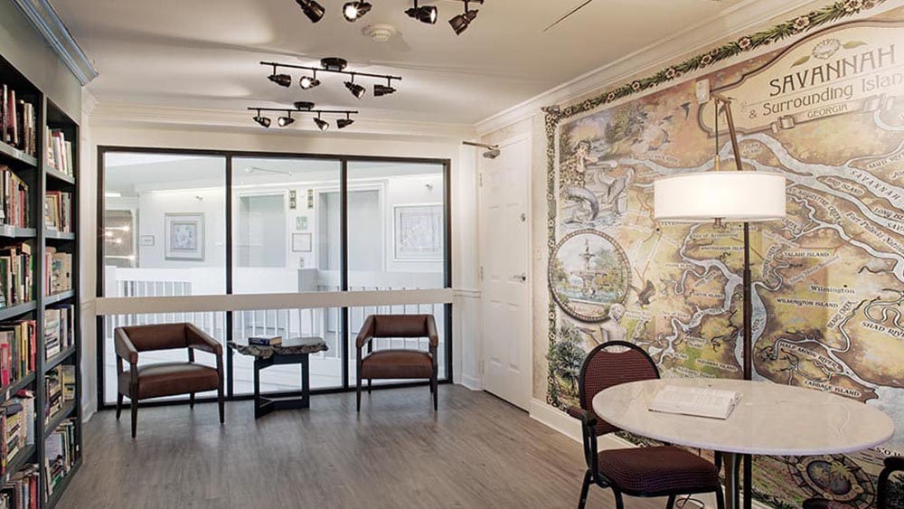 A small reading area with bookshelves, two chairs, and a round table, featuring a large illustrated map of Savannah on the wall and modern overhead lighting—perfect for relaxing at Thrive Senior Living.