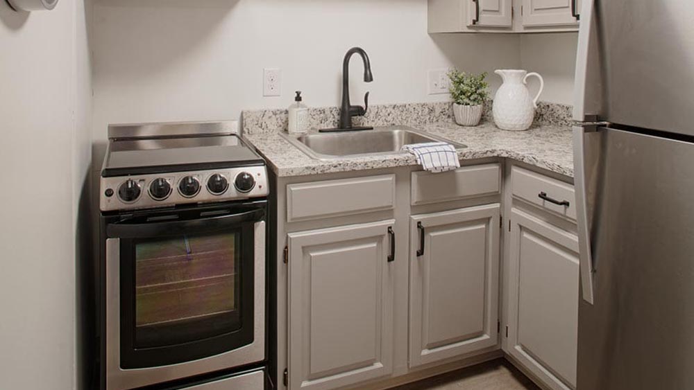 A compact kitchen at Thrive Senior Living features light gray cabinets, stainless steel appliances, a granite countertop, sink with black faucet, and minimal decor including a plant, soap dispenser, and pitcher.