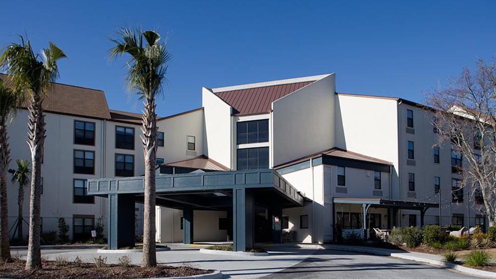 A modern, multi-story Thrive Senior Living building with a covered entrance, palm trees, and a clear blue sky in the background.
