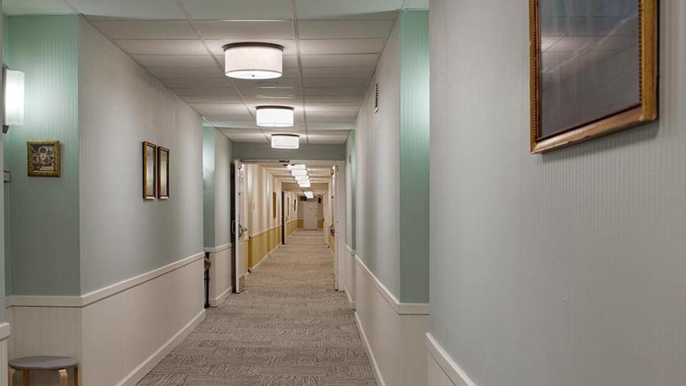 A long, well-lit hallway at Thrive Senior Living features light blue and white walls, framed pictures, and gray carpet flooring. Ceiling lights line the corridor, creating a welcoming atmosphere.