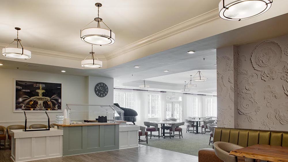 A spacious, well-lit dining area at Thrive Senior Living features modern light fixtures, tables and chairs, a clock on the wall, and large windows letting in natural light.