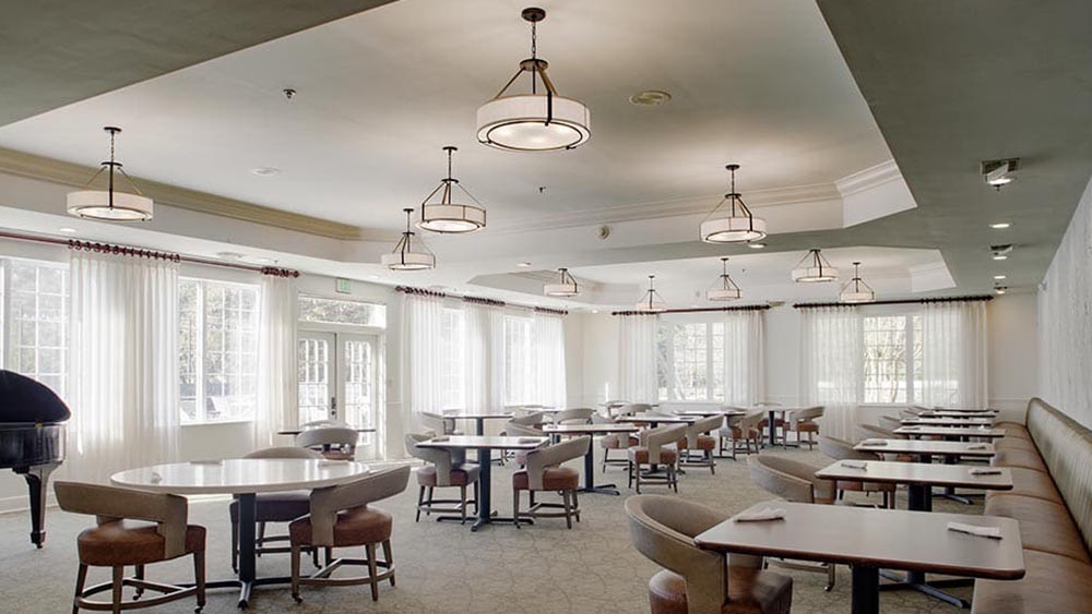 Spacious Thrive Senior Living dining room with round and rectangular tables, cushioned chairs, booth seating, large windows, white curtains, and ceiling light fixtures.