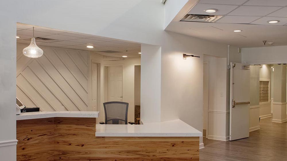 Modern reception area at Thrive Senior Living with a wooden front desk, white countertops, a mesh office chair, and geometric wall paneling, all under recessed lighting in a clean, well-lit space.