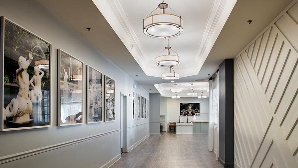 A well-lit hallway at Thrive Senior Living features modern light fixtures, white walls, large framed artwork on the left, and a patterned accent wall on the right, all leading to a welcoming seating area.