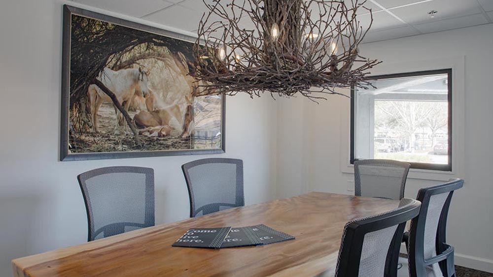 A conference room at Thrive Senior Living features a wooden table, four chairs, a twig chandelier, a window, and a large framed horse photograph on the wall.