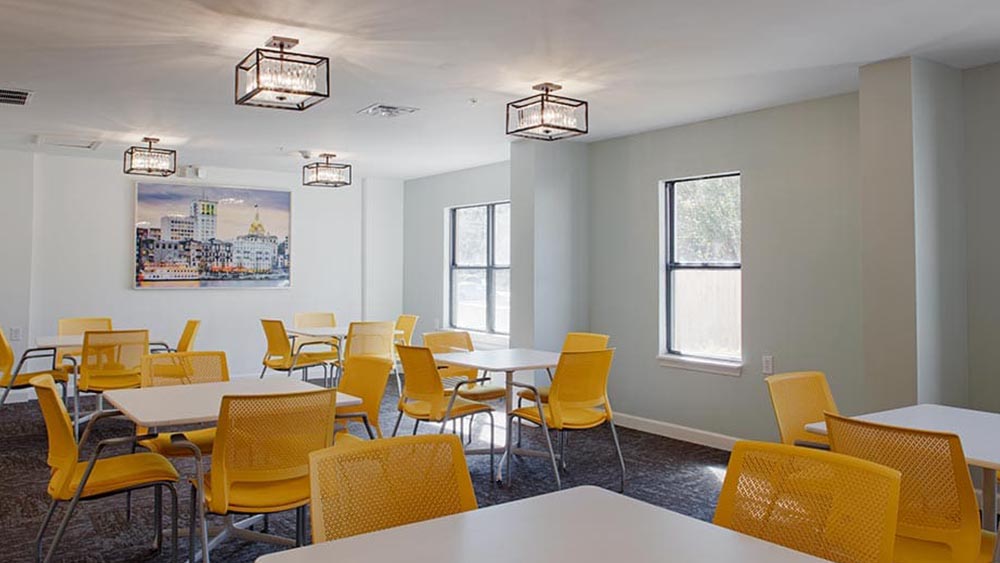 A modern room at Thrive Senior Living with several white tables and yellow chairs, gray carpet, light green walls, windows, ceiling lights, and a cityscape picture on the wall.