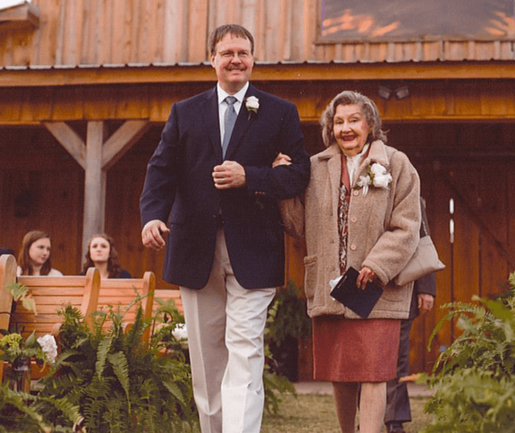 A man in a dark blazer and light trousers escorts an elderly woman, perhaps a Thrive Senior Living resident, down an outdoor aisle, with wooden benches and a building in the background.