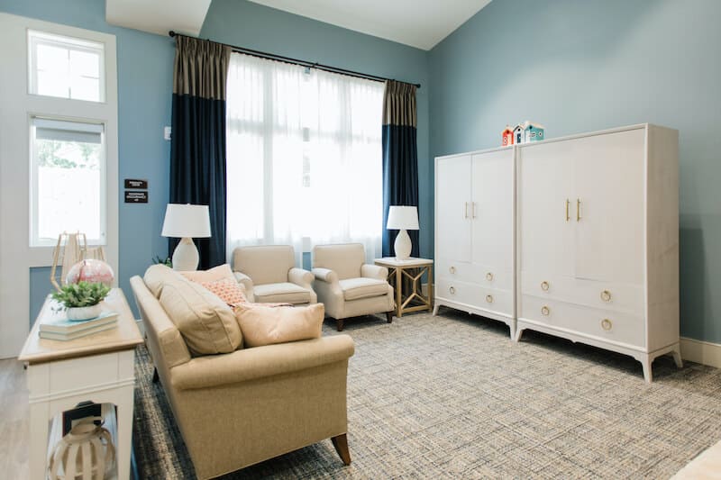 A living room at Thrive Senior Living features a beige sofa, two armchairs, white wardrobes, side tables with lamps, and a window adorned with sheer white curtains and dark drapes.