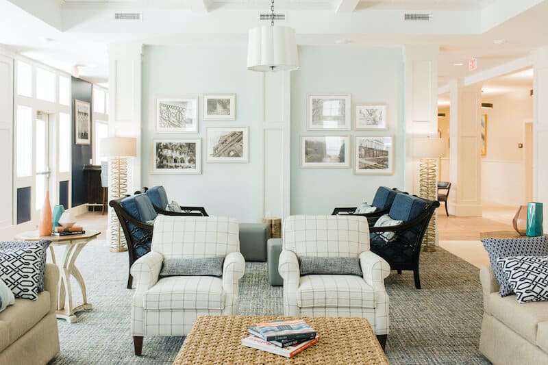 A bright, modern living room at Thrive Senior Living features two plaid armchairs, a woven coffee table, wall art, and inviting seating areas with blue cushions and throw pillows.