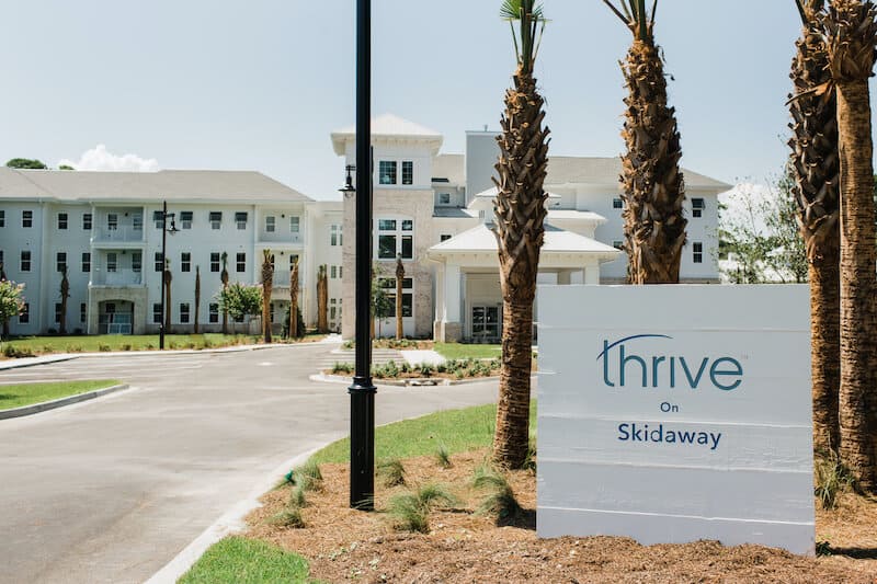 A modern, multi-story building with palm trees in front and a sign reading "Thrive on Skidaway" near the driveway entrance, showcasing the welcoming atmosphere of Thrive Senior Living.