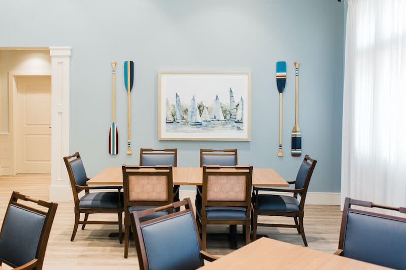 A dining area at Thrive Senior Living features wooden tables and blue cushioned chairs, nautical decor on pale blue walls, and a painting of sailboats between two decorative oars.