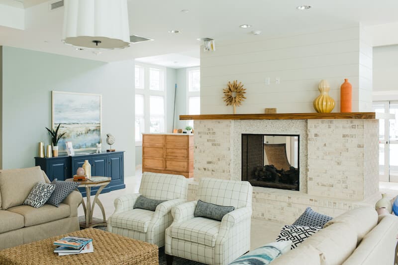 A bright living room at Thrive Senior Living features sofas, armchairs, a brick fireplace, decorative vases, a wicker coffee table, and a blue sideboard beneath a landscape painting.