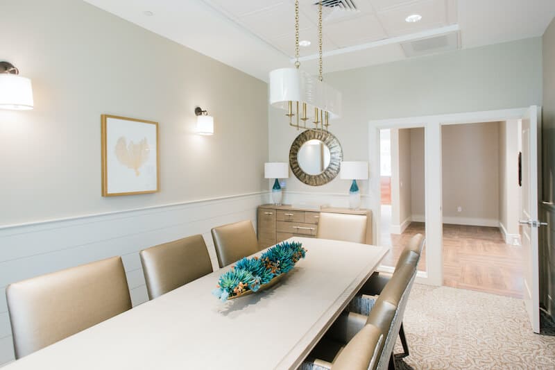 Modern dining room at Thrive Senior Living with a long table, six chairs, a decorative centerpiece, wall art, a mirror, and neutral-toned walls.