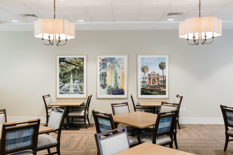 A dining area at Thrive Senior Living features square tables and chairs, three framed pictures on the wall, and two ceiling lights with beige shades.