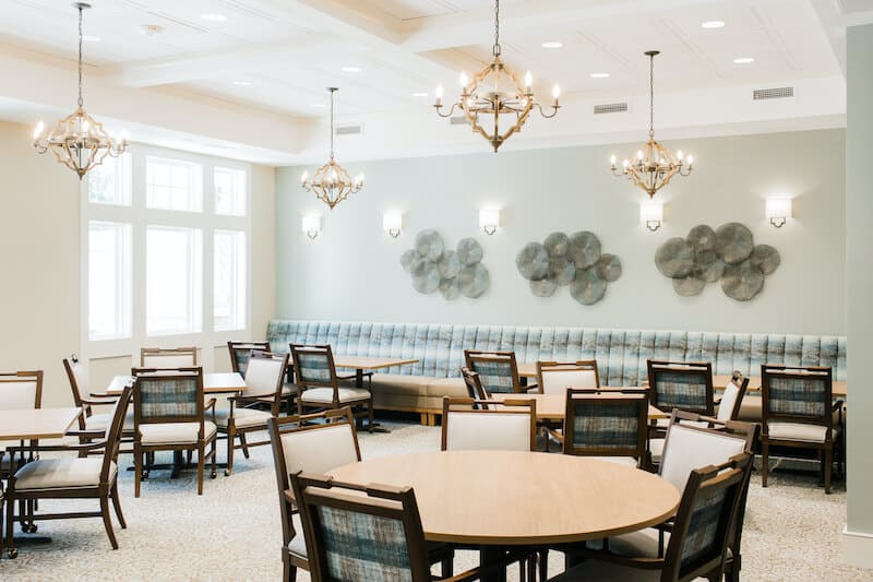 A bright, modern dining room at Thrive Senior Living features round tables, wooden chairs, upholstered bench seating along the wall, and decorative wall art under elegant chandeliers.