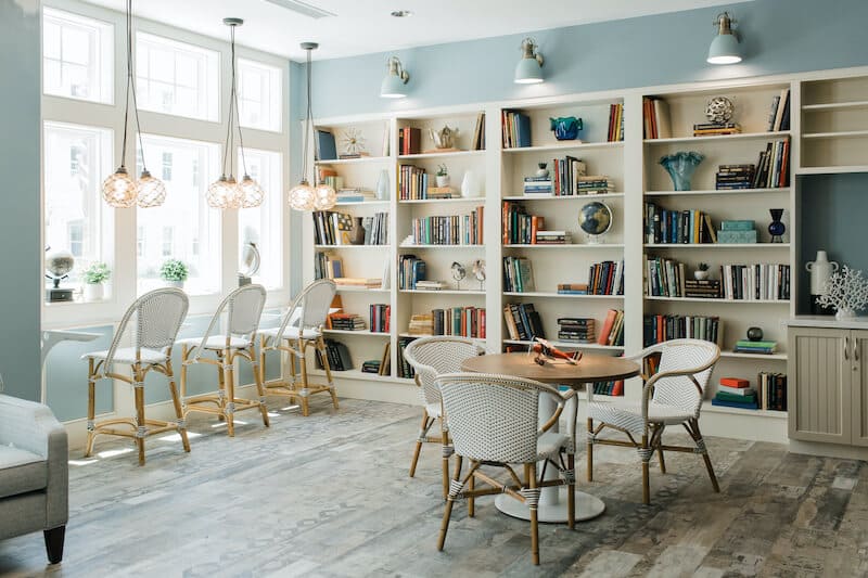 Spacious, well-lit room at Thrive Senior Living with bookshelves, a round table and chairs, plus a window counter with high stools for reading or working.