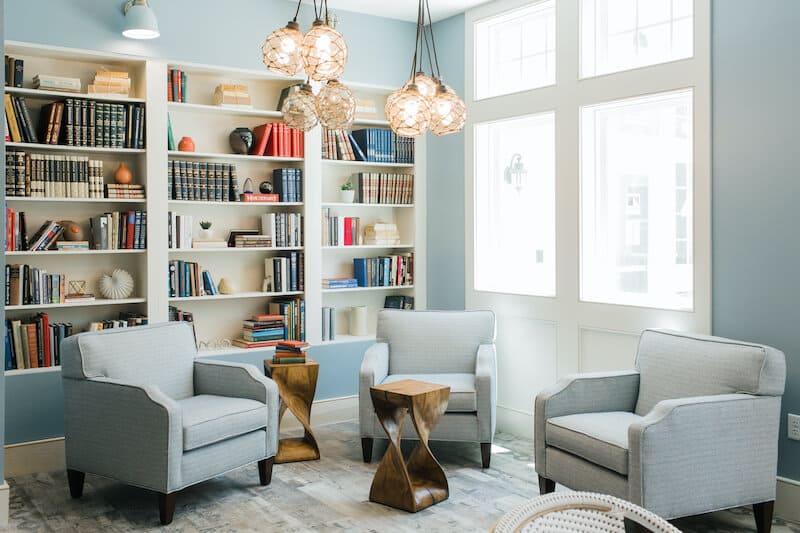 Modern reading room at Thrive Senior Living features three light gray armchairs, two wooden side tables, wall-mounted bookshelves filled with books, and large windows letting in natural light.